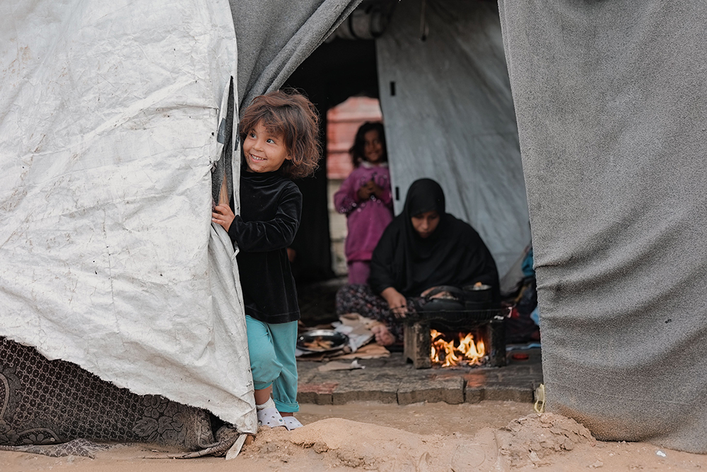 Young Palestinian girl smiling from the entrance of her family's tent in Gaza, illustrating the resilience of children living amid displacement.