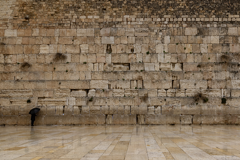 A lone person stands before the Western Wall in Jerusalem beneath an overcast sky.